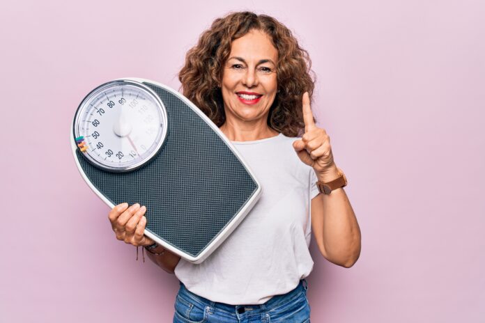 Middle age beautiful woman controlling weight using weighting machine over pink background smiling with an idea or question pointing finger with happy face, number one